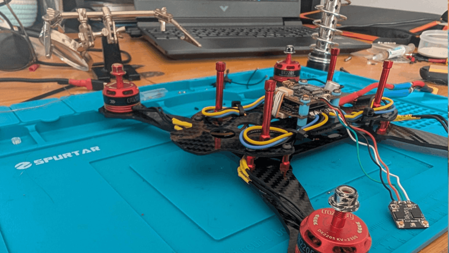 Deconstructed FPV drone components laid out on workshop bench with soldering tools and assembly equipment.